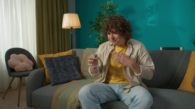 Young man in casual clothing, sitting on the sofa in the living room drinking water and choking