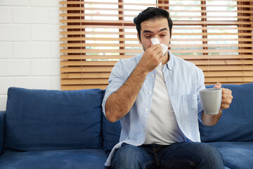 young man having a cold, using tissue paper and blowing his nose on sofa