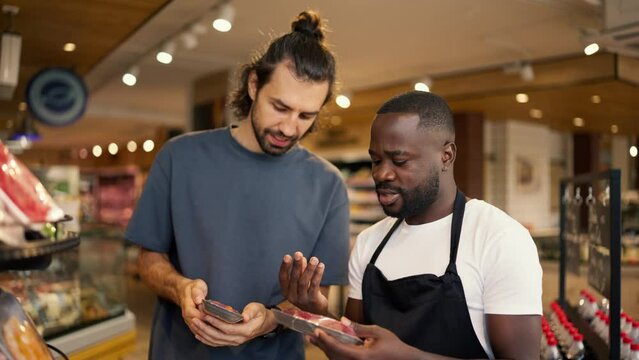 a man with Black skin color in a white t-shirt and a black apron helps a customer of a supermarket to choose a product
