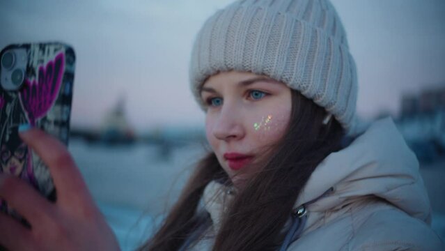 A young caucasian girl with red lipstick, blue eyes and glitter on her face in a winter jacket, long hair and a white hat takes pictures of winter sea and hills on smartphone with multi-colored case