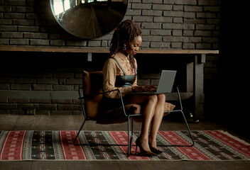 Attractive black woman using her laptop while sitting in a chair at the stylish interior with black brick walls and ornament rug. Working, studying, doing internet shopping, checking social networks