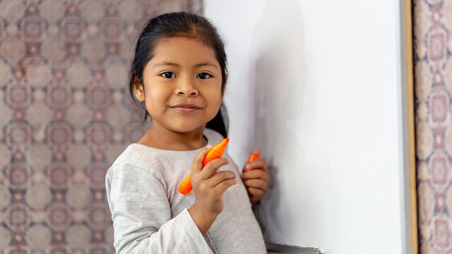 Ni&ntilde;a latina sonriente y feliz, escribiendo en la pizarra de clases con plumones de colores. Espacio para texto.