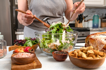 female hands make caesar salad with vegetables and chicken in a glass bowl. Rusks, chicken and salt in the foreground.