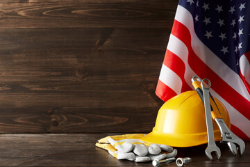 Salute the unwavering commitment of construction professionals this Labor Day. Side view photo of USA flag, wrenches, work helmet, gloves on brown wooden background with empty space for ads or text