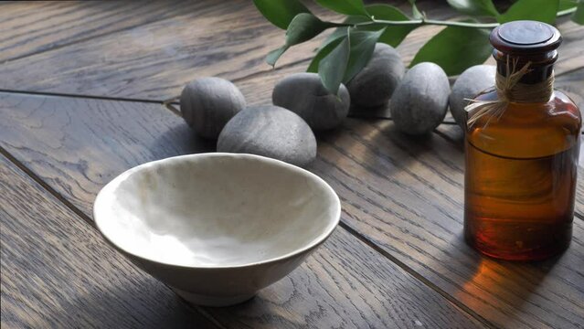 Base oil pouring from bottle to bowl on wooden background with stones and leaves