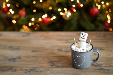 A marshmallow man sits in a mug of cocoa on the background of a Christmas tree. Merry Christmas