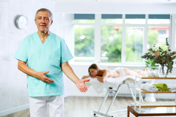 Smiling elderly masseur standing in massage cabinet with his back to patient waiting for procedure
