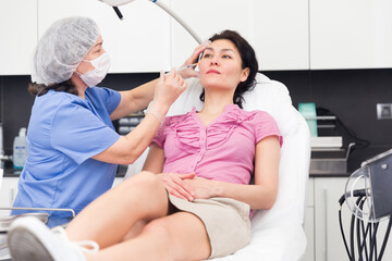 Focused female cosmetologist in a protective mask, working in the clinic, gives a mesotherapy injection to a young ..woman patient lying on the couch