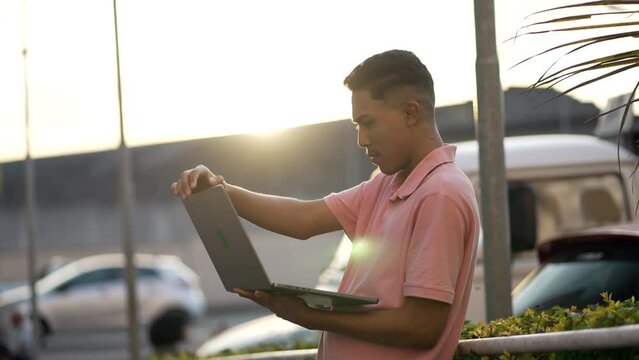 One young hispanic man standing outside opening laptop screen working remotely. South American latin student stands outdoors using modern computer during sunset