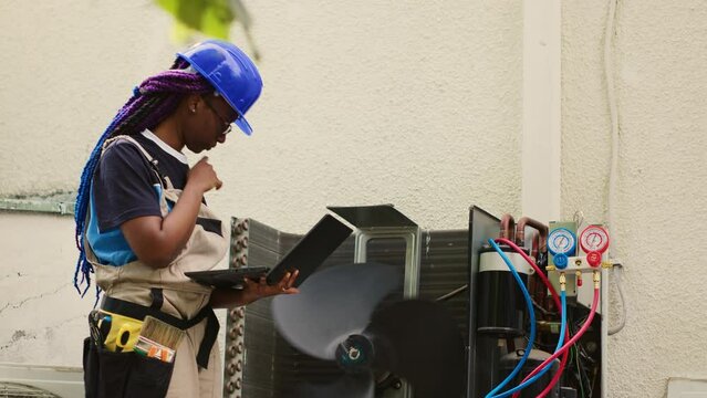 Seasoned mechanic servicing outside HVAC system internal parts, checking freon level and calibrating thermostats. Adept repairman imputing maintenance results data on laptop