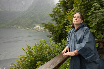 Fototapeta premium A young woman with a birthmark stands against the background of summer greenery and a lake on a rainy day in a raincoat