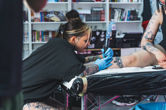 Busy day at tattooing studio. Female artist sitting on a chair and using professional tattooing equipment to draw a leg tattoo. High quality photo