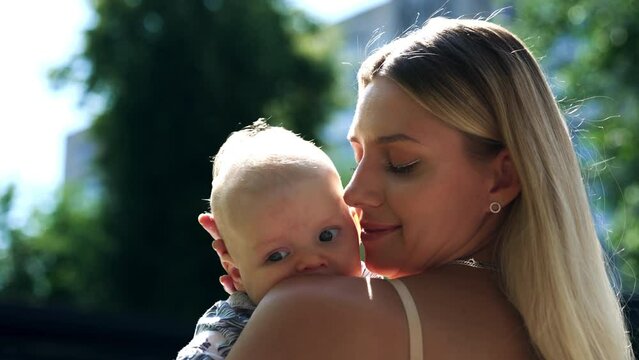 Beautiful blonde woman stands half-back to camera holding a baby. Loving mom caressing her son tenderly. Close up.