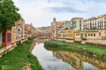 Fototapeta na wymiar Colorful yellow and orange houses and bridge Pont de Sant Agusti reflected in water river Onyar, in Girona, Catalonia, Spain. Church of Sant Feliu and Saint Mary Cathedral at background. BRIDGE in the