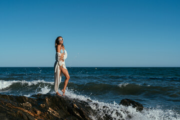 A beautiful girl in white glasses and white clothes sits on the rocks on the seashore