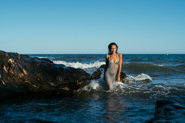 A beautiful wet brunette girl in a silvery net stands in the water on the seashore