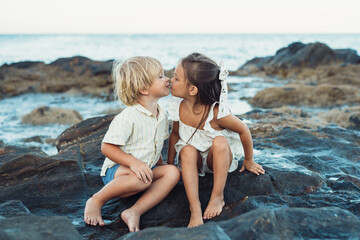 a blond boy and a brunette girl three years old are kissing while sitting on stones on the seashore