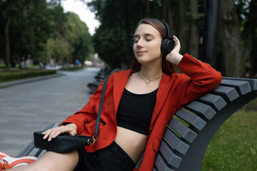 Happy girl in stylish clothes sits with her eyes closed on park bench and listens to music on headphones. Generation Z. Relaxation, rest, romantic mood
