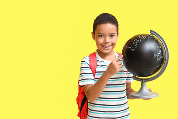 Little African-American boy with school backpack pointing at globe on yellow background