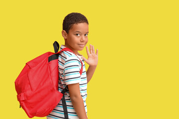Little African-American boy with school backpack waving hand on yellow background