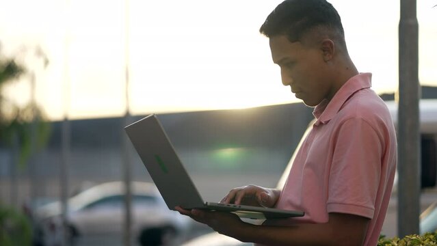 South American young man standing outdoors in front of laptop screen during sunset time. A hispanic Brazilian latin person working remotely in front of computer