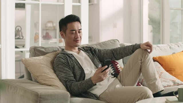 Young Asian Man Sitting Lying On Sofa At Home Looking At Cellphone