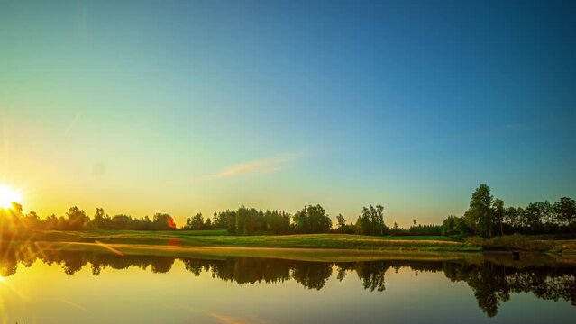 Cinematic timelapse of clouds moving fast over the sky before the sun rises on a bright day over a lake with trees and grass reflecting in the calm waters, Time Lapse