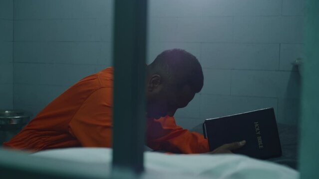 Religious African American prisoner in orange uniform kneels near the bed, prays to God holding Bible. Prison cell with barred window. Male criminal in jail or correctional facility. Faith in God.
