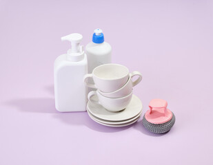 Obraz na płótnie Canvas Clean white plates and cups, detergents and an abrasive dishwashing sponge on a purple background.
