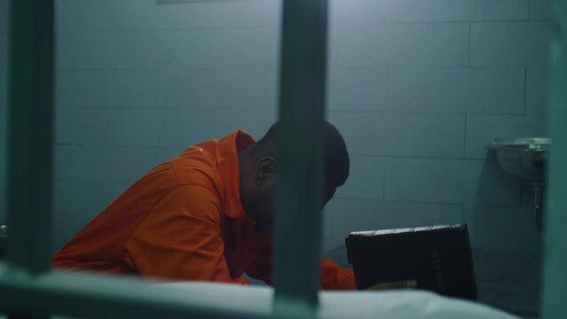 Religious African American prisoner in orange uniform kneels near the bed, prays to God holding Bible. Male criminal looks at barred window in prison cell. Jail or detention center. Faith in God.