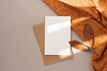 Blank paper card mock up, envelope, brown fall leaves, orange knitted textile on neutral beige...