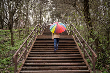 young woman in a beige coat and rain boots stands in forest or park and holds colorful bright...