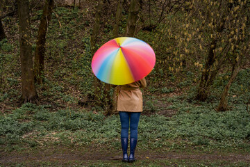 young woman in a beige coat and rain boots stands in forest or park and holds colorful bright rainbow umbrella. Girl spins umbrella for blur effect. Slow shutter speed. Autumn weather in city