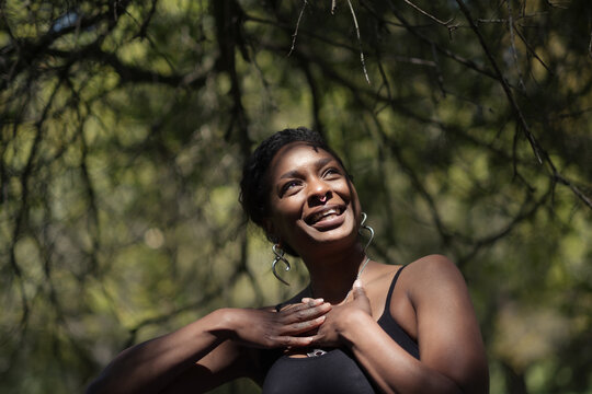 A person holds their hands on their chest while looking up towards the sunlight with a beaming smile. Behind them are shadowy, mysterious tree branches. The photo expresses hope and optimism.