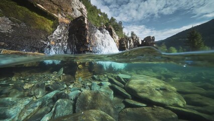 A shallow river with rocky shores and transparent waters, with a waterfall in the background. Over-under water shot.