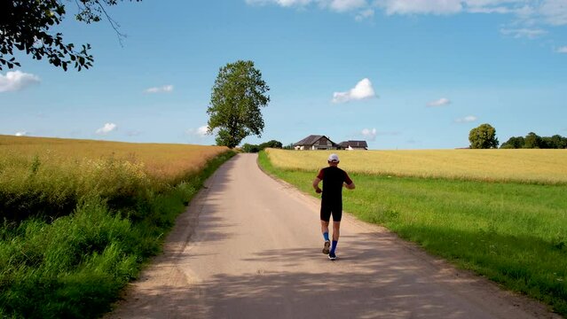 Following a running man closely from behind - a deserted asphalt path between ripening fields and the embrace of nature - drone shot.