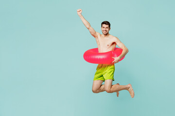 Full body side view young man wear green shorts swimsuit relax near hotel pool jump high in rubber ring do winner gesture isolated on plain blue background. Summer vacation sea rest sun tan concept.