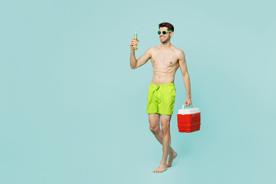 Full body side view young man wears green shorts swimsuit relax near hotel pool walk go with fridge box hold bottle of beer isolated on plain blue background. Summer vacation sea rest sun tan concept.