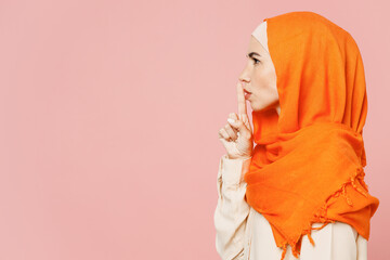 Side view young arabian asian muslim woman wearing orange abaya hijab say hush be quiet with finger on lips shhh gesture isolated on plain pink background. Uae middle eastern islam religious concept. - obrazy, fototapety, plakaty