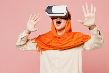 Young surprised arabian asian muslim woman wear orange abaya hijab watching in vr headset pc gadget isolated on plain light pink background studio portrait. Uae middle eastern islam religious concept.