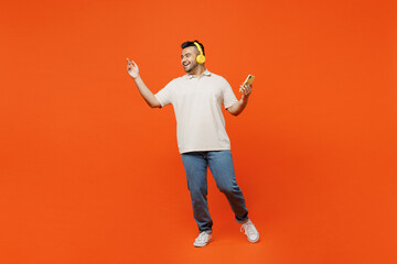 Full body happy young Indian man he wearing white t-shirt casual clothes listen in headphones using mobile cell phone dance isolated on plain orange red background studio portrait. Lifestyle concept.