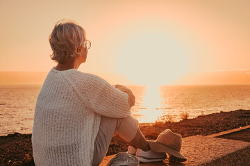 Back view of elderly mature woman sitting alone face the sea at sunset light looking at the horizon. Senior gray-haired woman enjoying freedom and beautiful seascape in vacation - Powered by Adobe