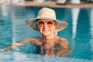 Portrait of smiling caucasian senior woman with hat and sunglasses relaxing into the outdoor swimming pool enjoying relax and vacation