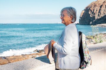 Smiling senior woman walking near the beach looking at the horizon enjoying beautiful seascape in vacation day - retired life