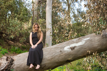 Young woman, blonde and beautiful, with a black dress, sitting on the trunk of a large tree, in the middle of nature, pure and virginal. Concept nature, peace, purity, virginity, trees.