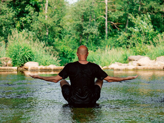 Bald man sitting in water in a stream doing yoga relaxing exercises. The model is bald wearing dark clothing. Doing workout in unusual exotic places. Fitness for mind and body. Active lifestyle