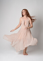 Full length portrait of beautiful  female model with long  brunette hair wearing a creamy pink gown dress. graceful dancing pose, with gestural hands  isolated on white studio background.
