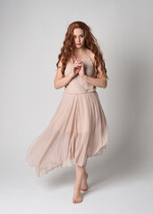 Naklejka premium Full length portrait of beautiful female model with long brunette hair wearing a creamy pink gown dress. graceful dancing pose, with gestural hands isolated on white studio background. 