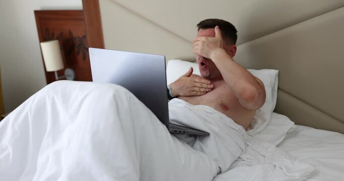Young man using laptop on bed scared and startled with open mouth in surprise. Distrustful face shocked internet news