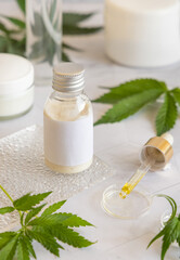 Cosmetic bottle with blank label near green cannabis leaves on white table. Mockup